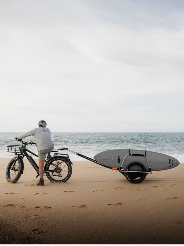 Une personne avec un vélo et une remorque Reacha sur une plage, transportant une planche de surf avec la mer en arrière-plan