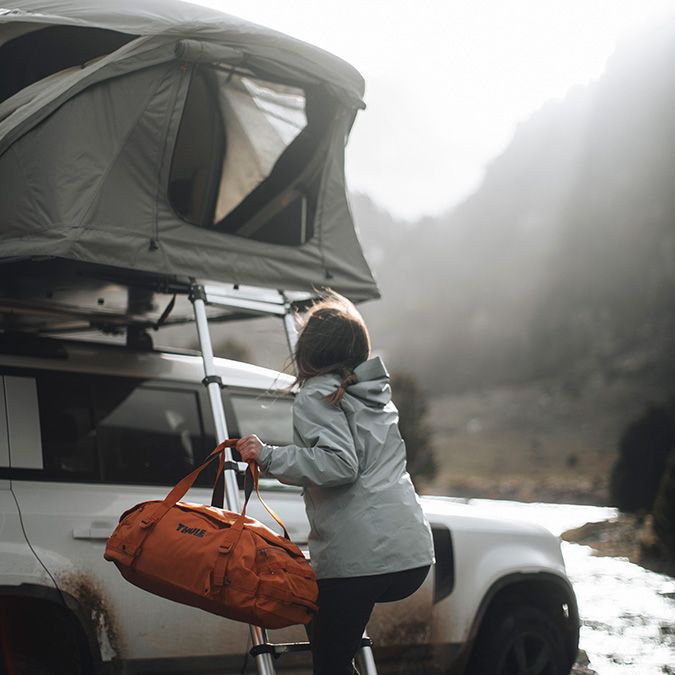 A woman climbs the ladder and enters her Thule Approach rooftop tent with a duffel bag