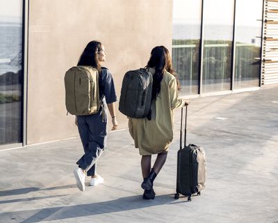 Two people walk down a street with travel backpacks and suitcases.