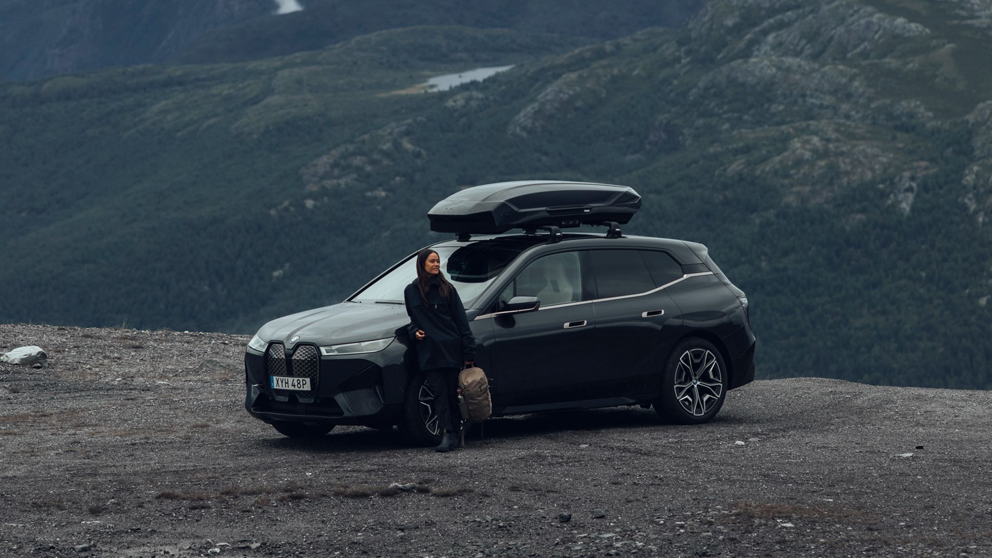 Una donna che tiene una borsa Thule si appoggia a un'auto con una scatola sul tetto Thule. 