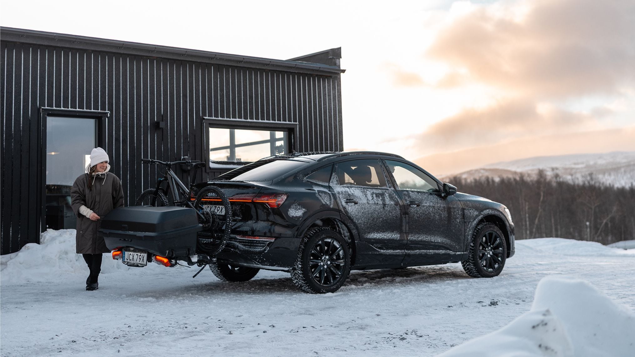 En mörk SUV med ett bakmonterat lastsystem som rymmer både en låda och cykel står parkerad bredvid en svart trähytt i ett vinterlandskap. En person närmar sig bakdelen av fordonet.