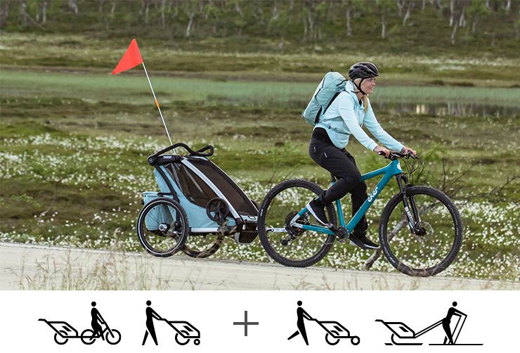 A woman bikes with a blue Thule Chariot Cross bike trailer through the mountainous countryside