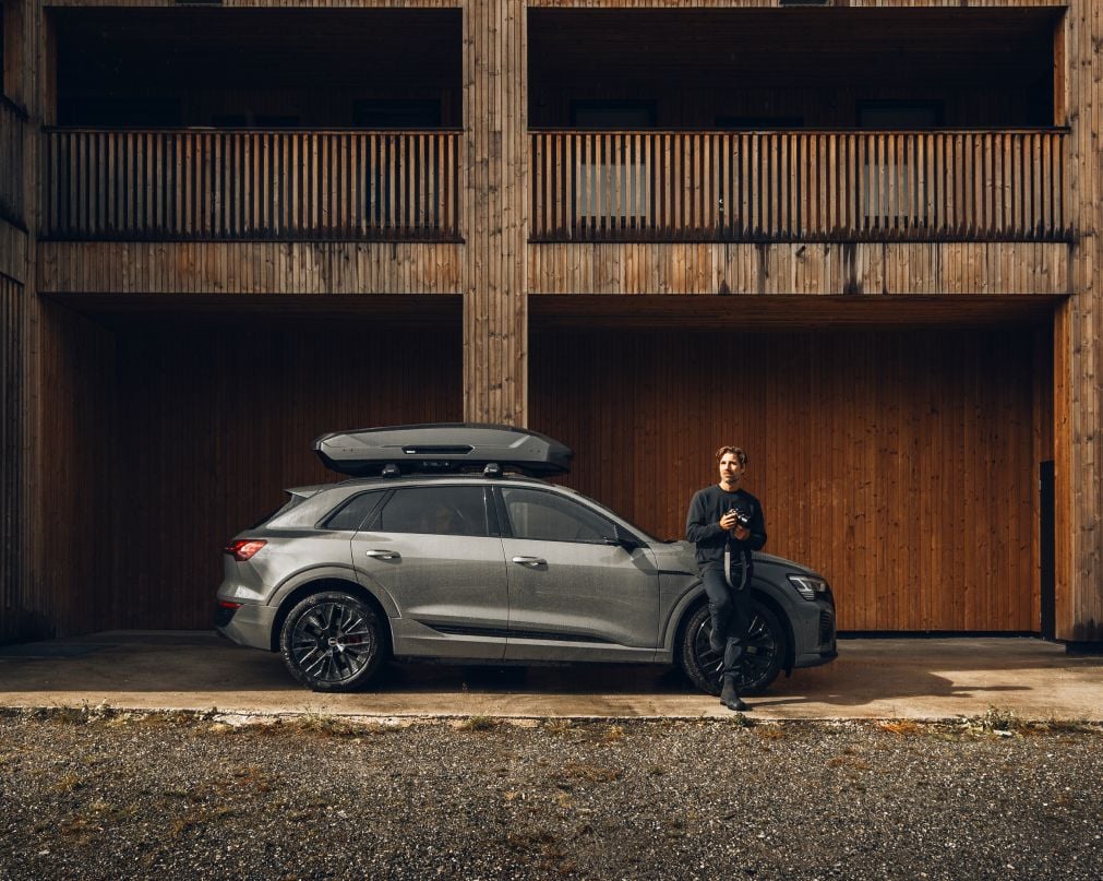 Un hombre apoyado en un automóvil de color gris con un baúl de techo Thule Motion 3.