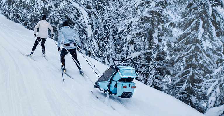 Пара поднимается на лыжах вверх по заснеженному холму, таща за собой ребенка в прицепе Thule Chariot Cross.