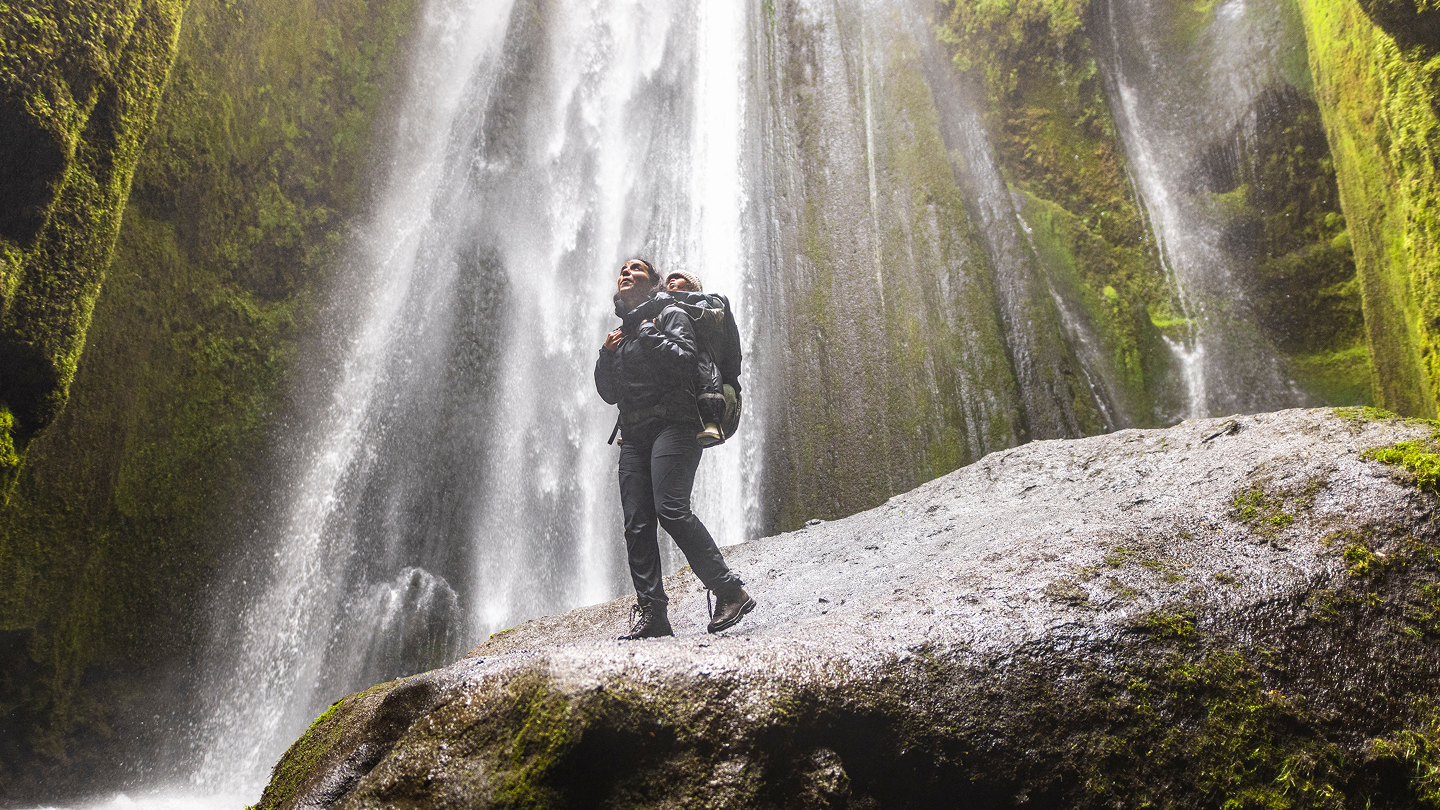 Una donna che indossa uno zaino marsupio Thule Sapling con un bambino dentro fa escursioni vicino alle cascate. 
