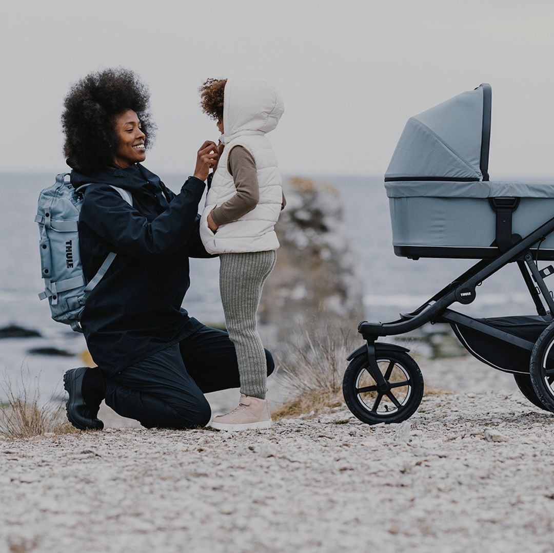Uma mulher usando uma mochila Thule segura um casaco infantil em uma praia ao lado de um carrinho Thule. 