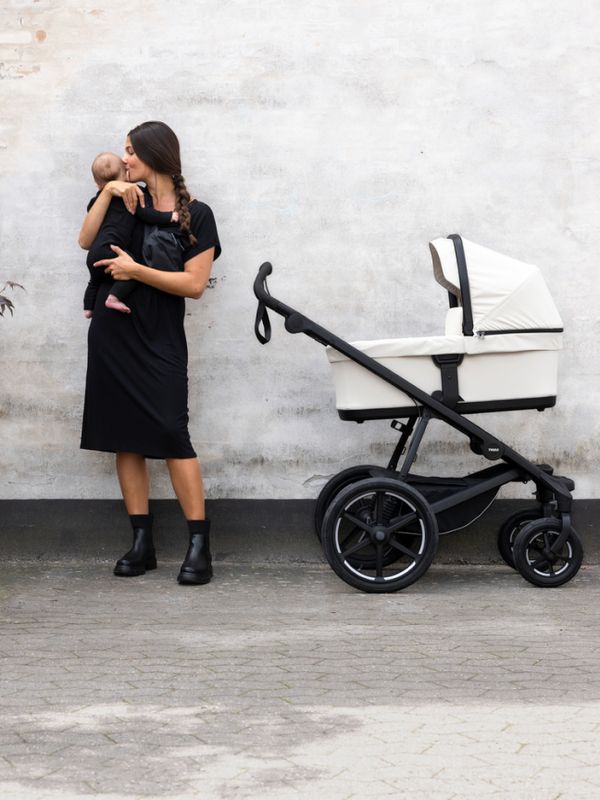 En mor i svart kjole står mot en minimalistisk betongvegg og holder babyen tett inntil seg mens en Thule Urban Glide 3-barnevogn står parkert ved siden av henne.