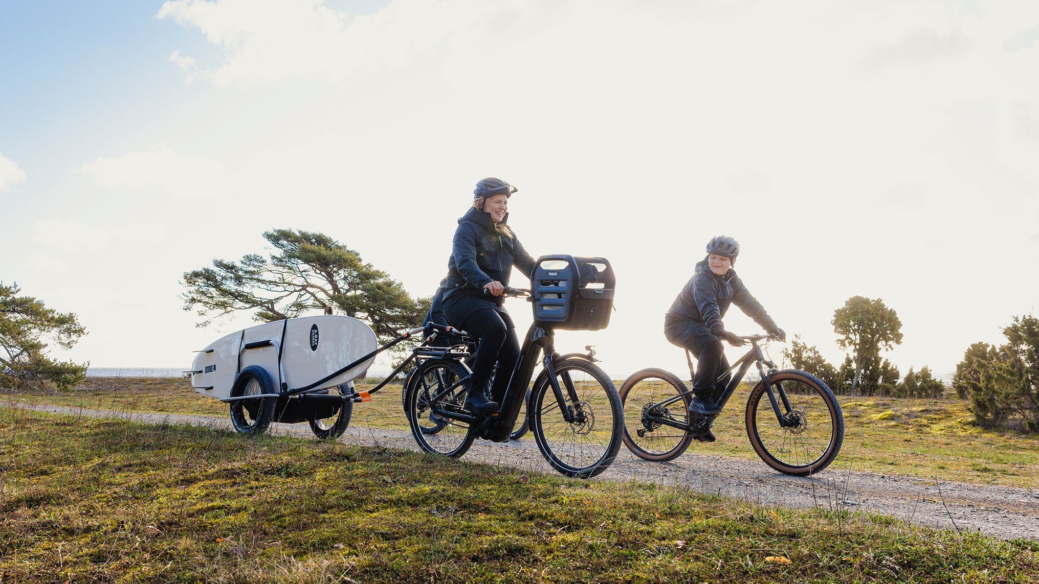Anja Pärson a pedalar com uma criança num caminho de cascalho, rebocando um atrelado de bicicleta Thule que transporta uma prancha de surf por uma paisagem costeira relvada.