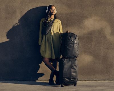 Женщина в наушниках стоит с чемоданом из коллекции Thule Aion в солнечную погоду.