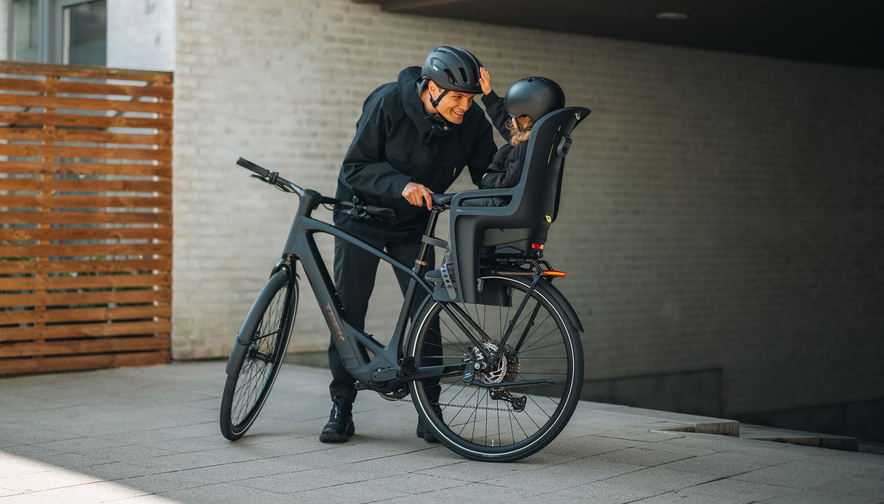 Adult leaning over a bicycle, interacting with a child seated in a Thule RideAlong 2 bike seat; both wearing helmets in a paved urban area.