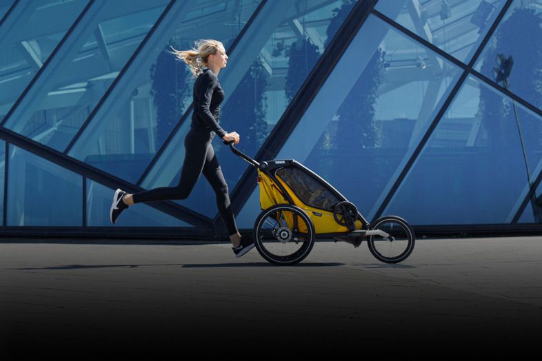 A woman runs past a modern glass building with her yellow Thule Chariot Cross jogging bike trailer.
