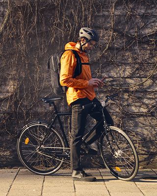 Мужчина в оранжевом и с велосипедным рюкзаком Thule Paramount едет на велосипеде по городской улице.