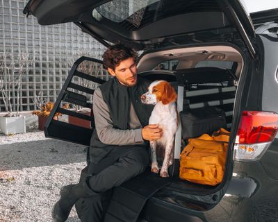 Un hombre está sentado en el maletero abierto de su coche junto con su perro y una jaula para perros Thule Allax en el fondo