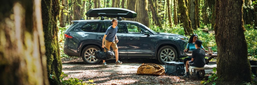 Cenário florestal com um carro equipado com Thule Roof Box e um homem na frente do carro. 