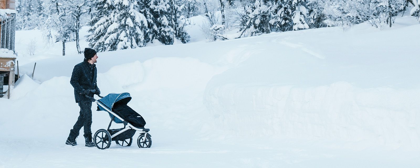 Mies kävelee lumessa joka maaston Thule Urban Glide 2 ‑lastenvaunujen ja mustan lämpöpussin kanssa.