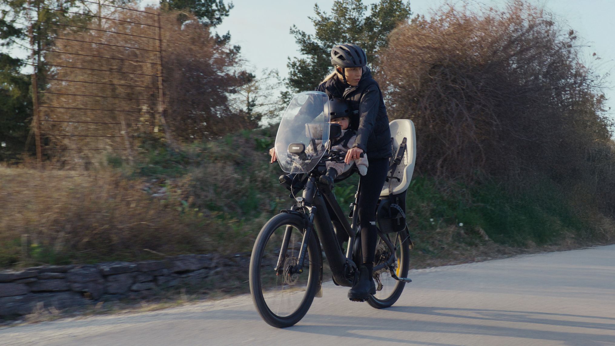 Kristin Larsson roule à vélo sur une route de campagne avec un enfant assis dans un siège vélo Thule à l’avant et un autre siège Thule monté derrière elle.