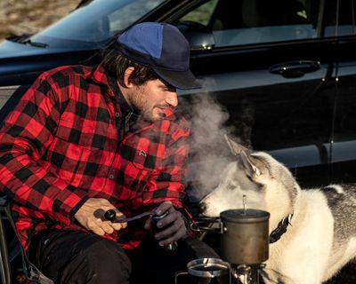 Muž ve venkovním oblečení se dívá na svého psa, zatímco připravuje jídlo na kempingovém sporáku.