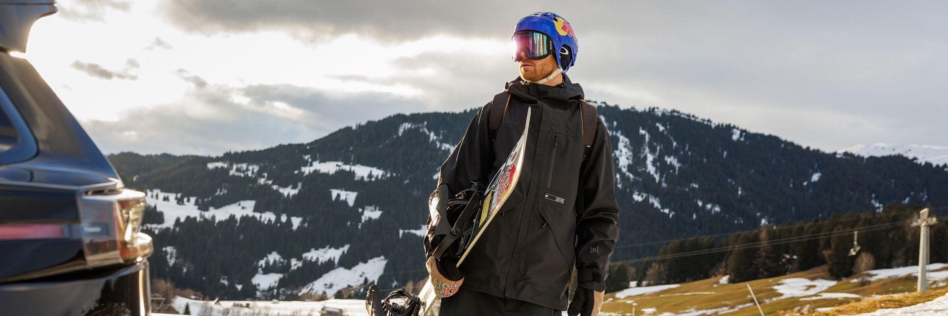En snowboardåkare som står utomhus i vinterkläder, håller i en snowboard och tittar mot bergen.