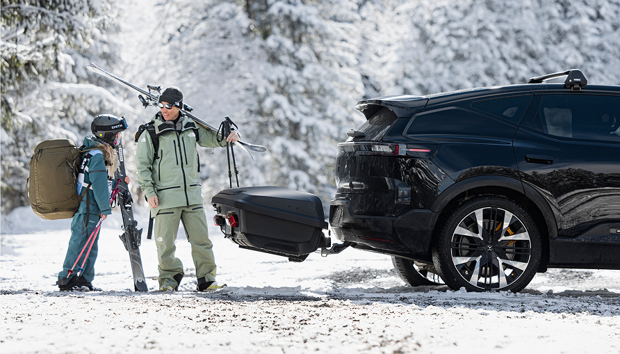 Two people standing beside a dark SUV with a rear-mounted cargo carrier in a snowy landscape, carrying skis and winter gear.