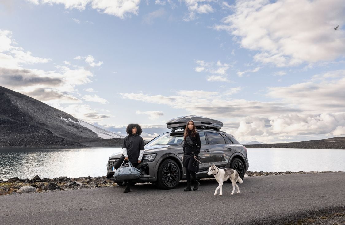 Duas mulheres e um cachorro estão parados na frente de um carro cinza estacionado perto de um lago. Um bagageiro de teto Thule Motion 3 está montado no carro. 