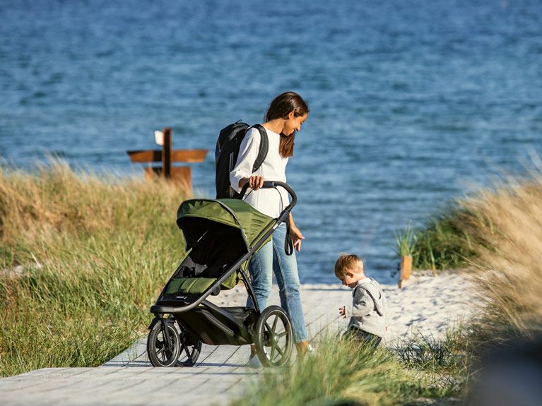 A woman watches a child play on the beach as she waits with a green all terrain stroller.
