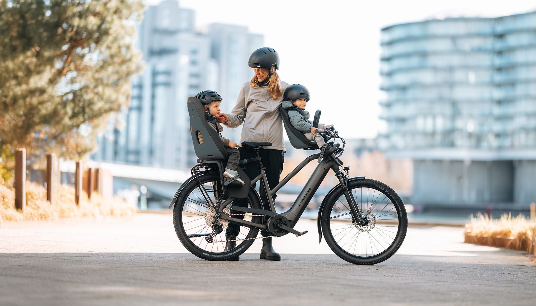 Adult standing with a bicycle fitted with two child seats, each carrying a child.