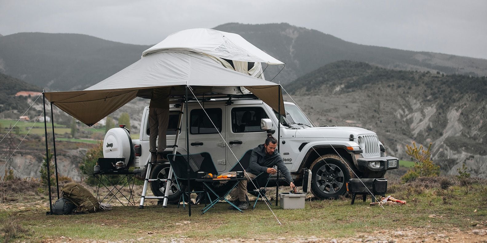 산에 Thule Approch 루프탑 텐트가 장착된 지프가 주차되어 있고 한 남자는 어닝 아래에 앉아 있습니다