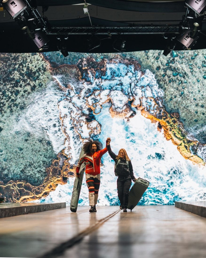 Two people performing on a runway in front of a large screen showing a vibrant aerial landscape.