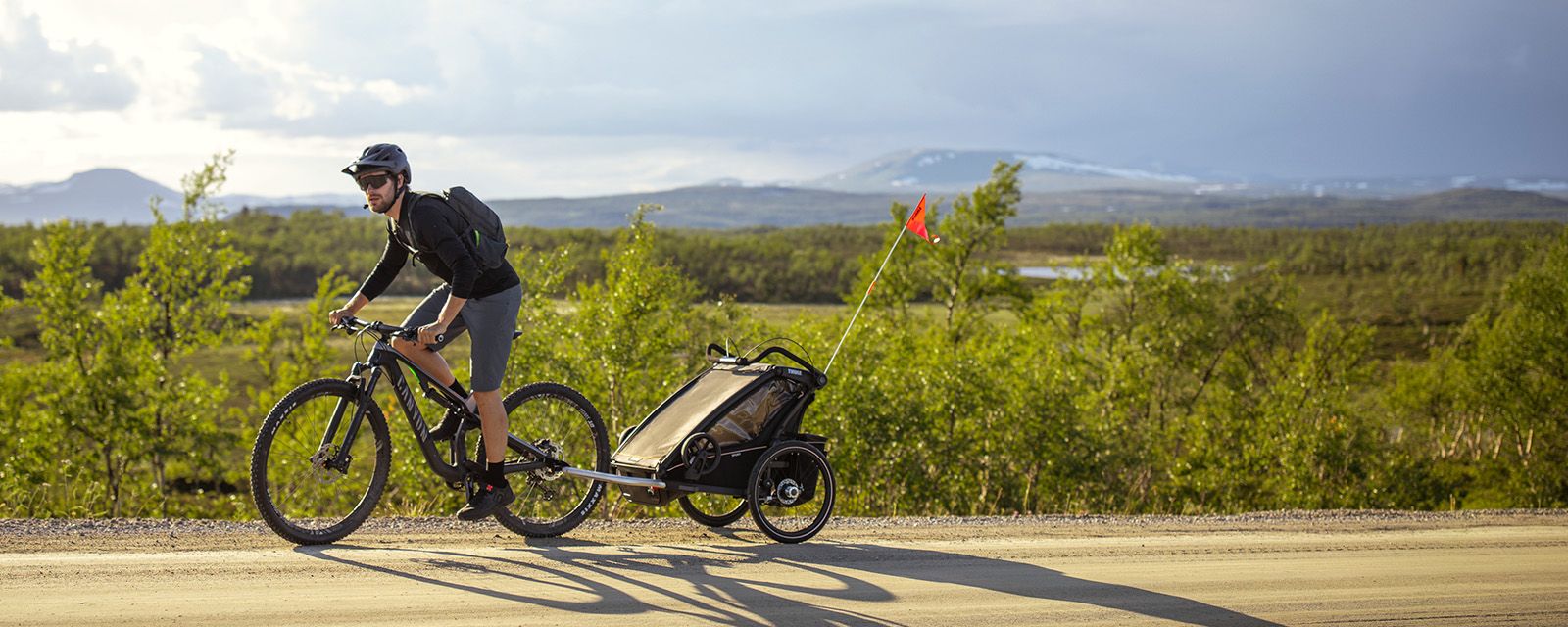 In an expansive grassland a man goes biking with a black Thule Chariot Sport child bike trailer.