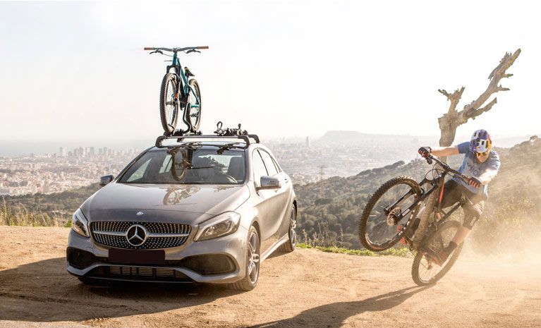 Mountain biker on a bike next to a car with a Thule roof bike rack with a city in the background.