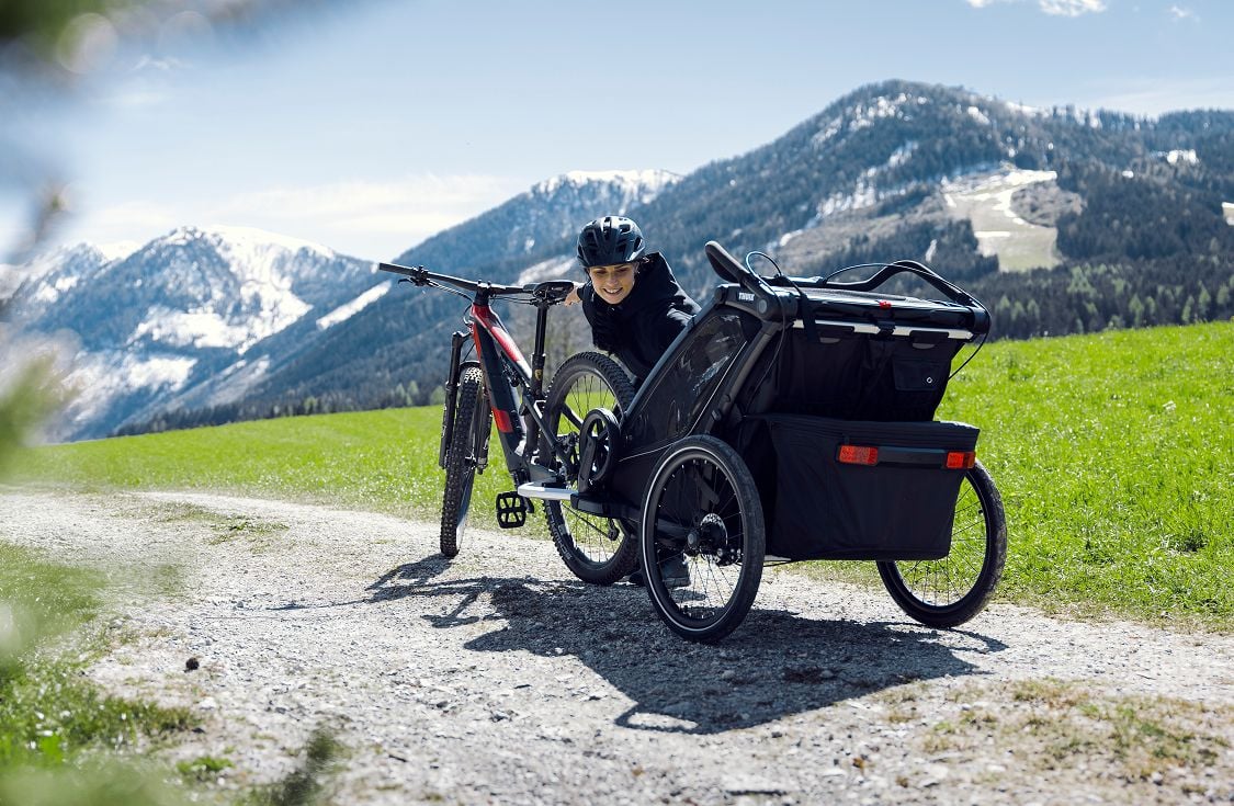 Knielende persoon naast een mountainbike met een fietsaanhanger op een grindweg in een berglandschap.