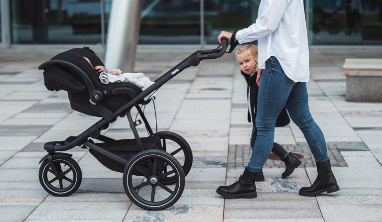 A woman walks with her baby in a Thule Urban Glide car seat stroller while her toddler walks beside her.