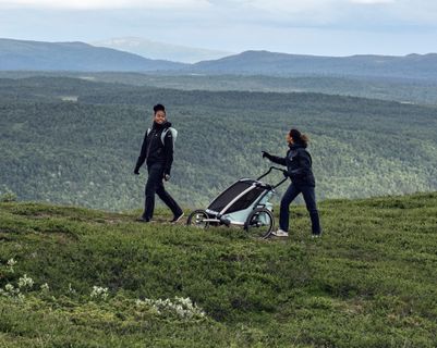 Two women wearing coats walk in the mountains while one pushes a Thule Chariot trailer on a path.