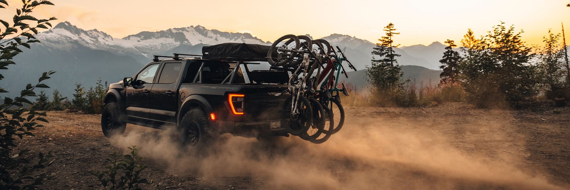 Pickup truck driving on a dusty mountain road at sunset with multiple bikes mounted on a Thule Revert bike rack, promoting Bring Your Bike.