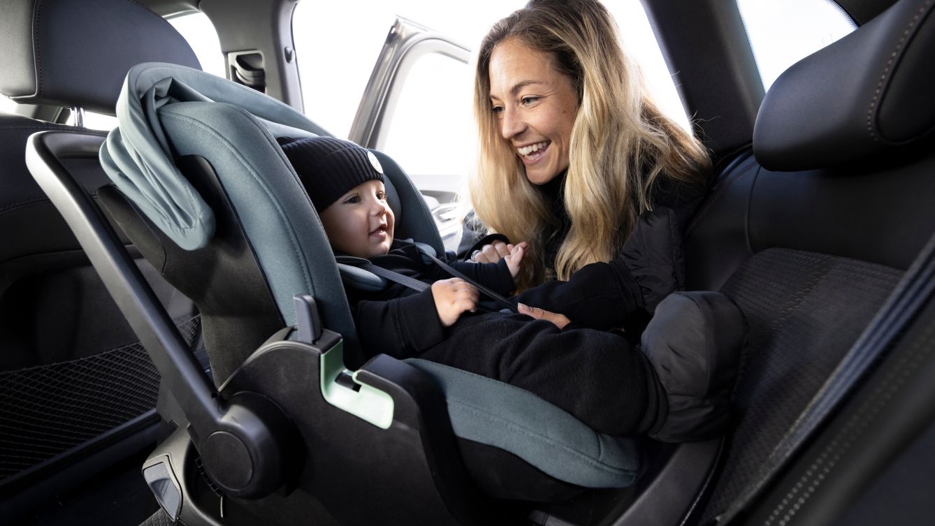Uma mulher com cabelo louro inclinada dentro de um carro e sorrindo. Um bebê está dentro do carro sentado no assento infantil para veículos voltado para trás Thule Maple.