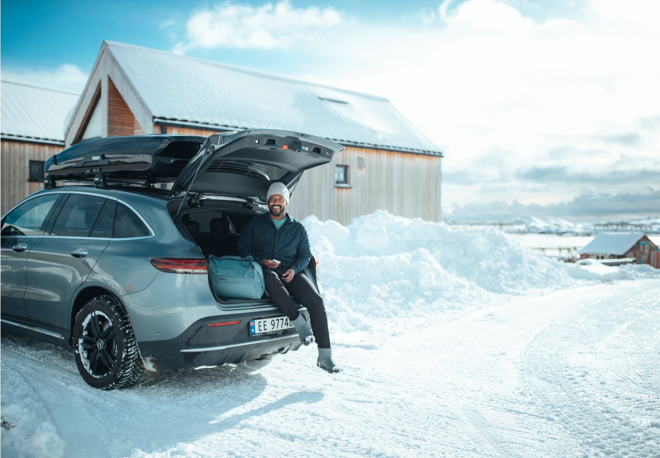 Mees istub auto pagasiruumis Thule Chasmi reisikoti kõrval. Auto katusel on Thule Motion 3 katuseboks ja see on pargitud lumisele alale. 