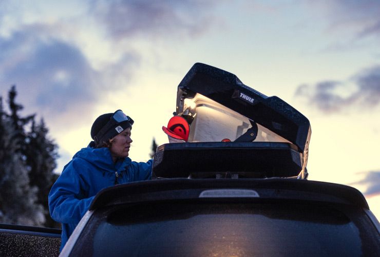 A close-up of a skier opening their Thule rooftop cargo carrier in a forest at sunset.