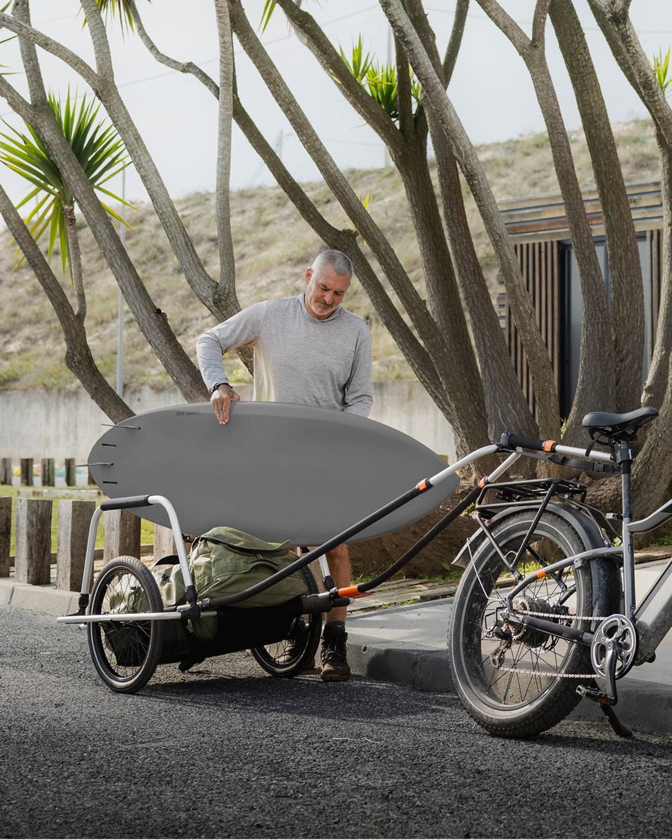 Erwachsener hebt ein großes Surfbrett von einem Fahrradanhänger, der an einem Fahrrad neben einem Bordstein geparkt ist.