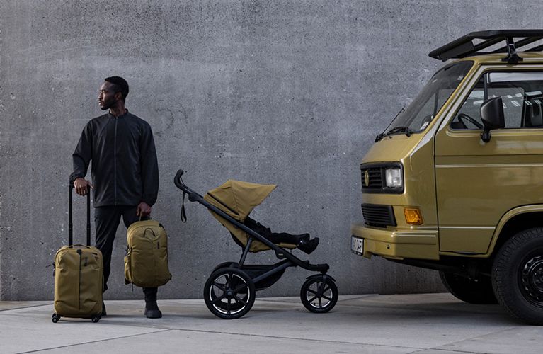 A man holding a piece of Thule luggage and a Thule backpacks stands next to a Thule Urban Glide 3 stroller.