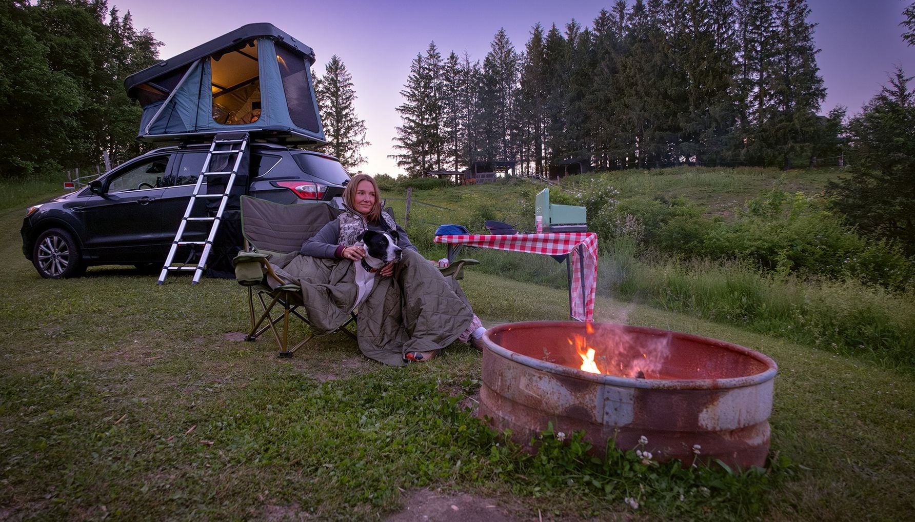 Woman  with a dog tucked beside her in a camp chair and an SUV with an open rooftop tent behind