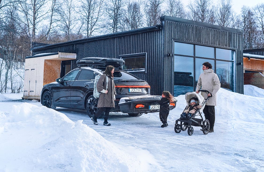 Uma família parada ao lado de um SUV escuro com uma cabine de proteção e um Thule Arcos de carga traseiro. Ele está estacionado em uma cabana moderna em uma paisagem nevada, com um adulto empurrando um carrinho e uma criança caminhando em direção ao veículo.