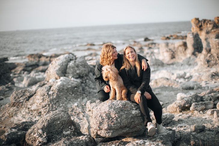 Duas mulheres sentadas com o seu cão numa praia rochosa.
