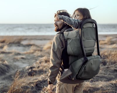 Un padre camina con su hija colocada en una mochila para transportar niños en la playa.