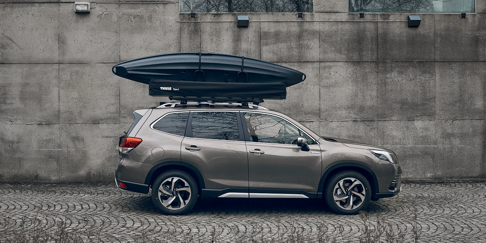 A vehicle parked next to a wall with a Thule Caprock roof platform, kayak and rooftop tent on the roof.