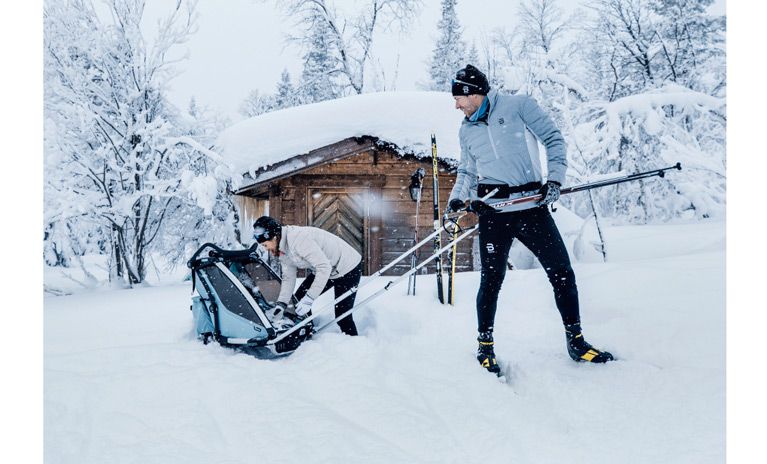 A couple in a snowy forest stand outside a wood cottage with their Thule Multi-sport cross-country ski bike trailer.