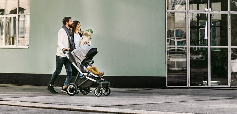 A couple walk with one of the Thule city strollers down a street next to a green building with a glass door.