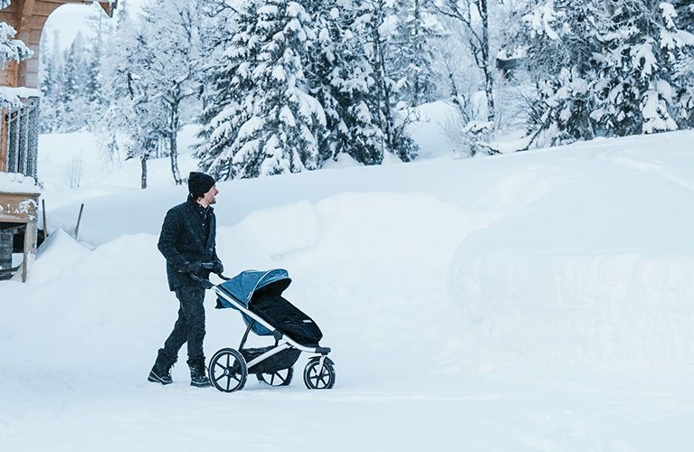 En man promenerar i snön med en Thule Urban Glide 2 joggingvagn för all terräng och en svart åkpåse.