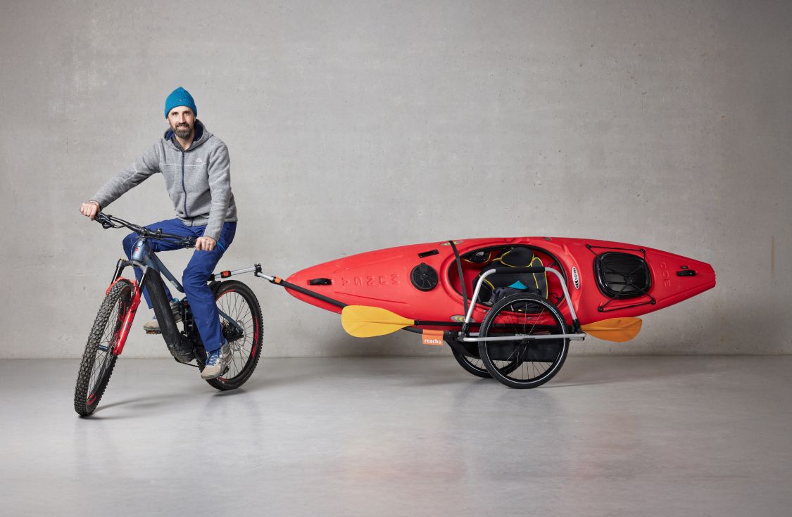 Un hombre montado en una bicicleta con un remolque Reacha Sport enganchado en el que lleva un kayak naranja intenso.