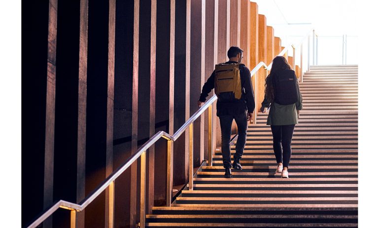 Two people walk up some stairs into the sunlight, wearing Thule Paramount backpacks.