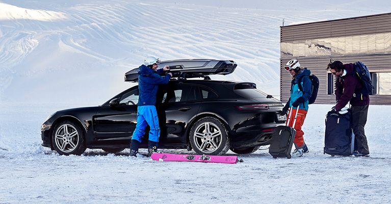 Два человека в лыжном снаряжении укладывают багаж в припаркованный в снегу автомобиль и в грузовой бокс на его крыше.